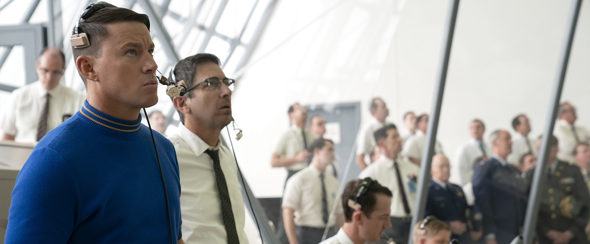 A group of men wearing shirts and ties are stood in a brightly lit room, some are wearing headsets and working at computers.