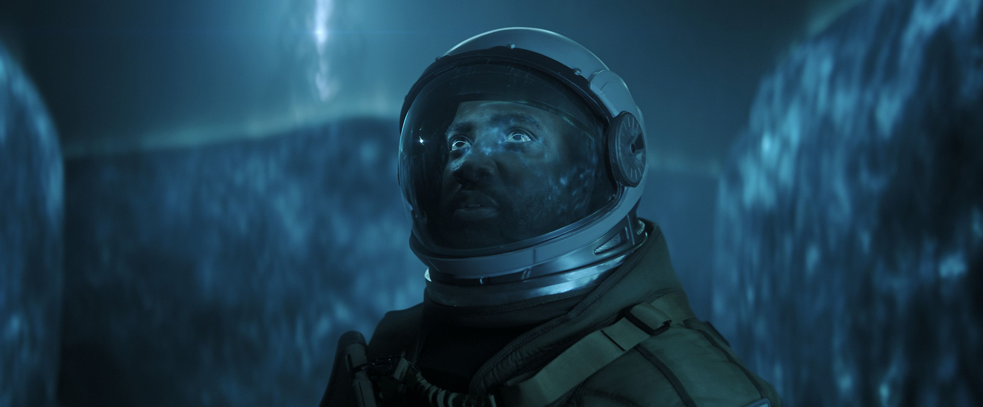 A person in a spacesuit stands in front of a neon blue extraterrestrial-like gaseous wall with an alien entity's silhouette looming  