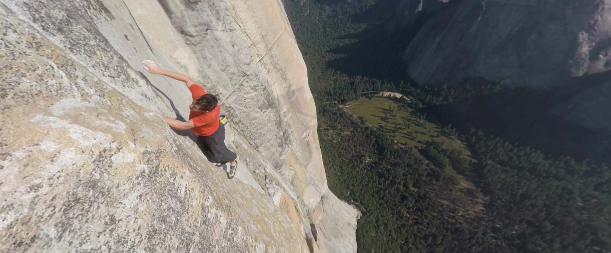 A man free climbing a large rock face above a forested hillside.