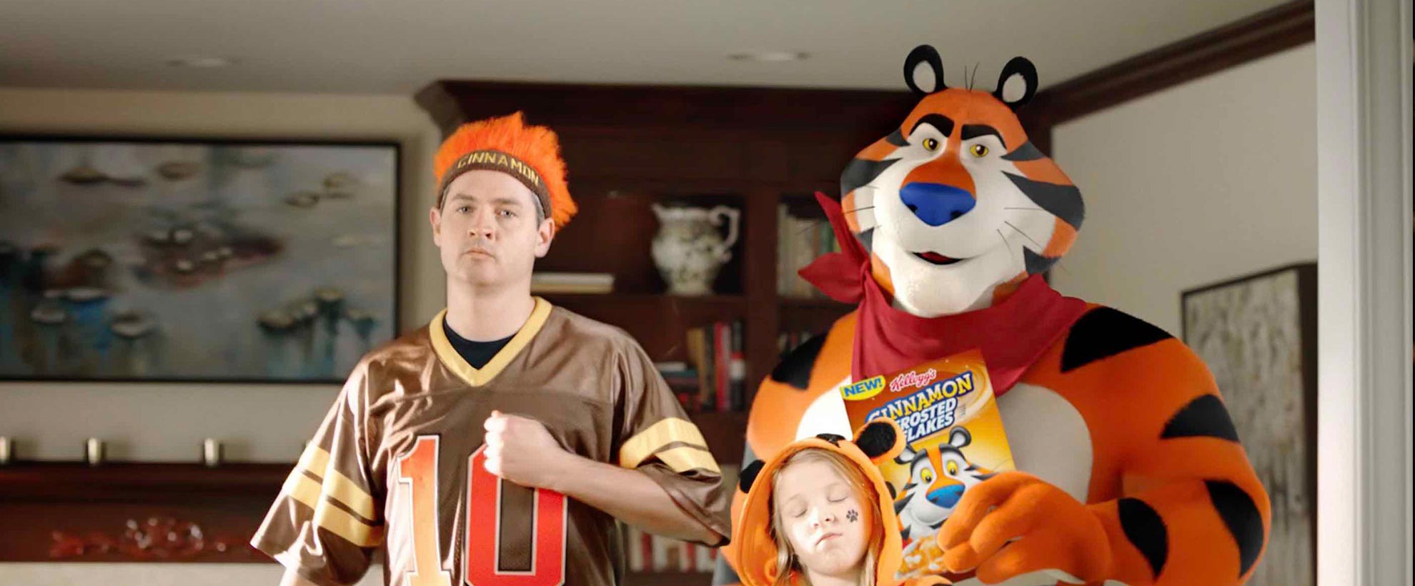 CGI Tony the tiger holds a box of frosted flakes cereal and stands next to a young girl in a tiger hood and a man in a sports jersey