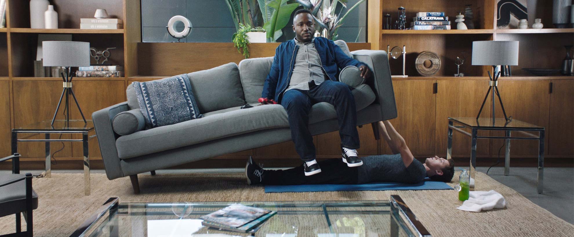 A man sits on a sofa which is being bench-pressed by another man lying underneath it