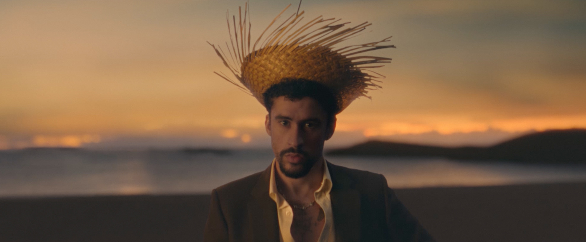 Bad Bunny wearing a spiky straw hat and an open shirt sits on a beach at sunset, looking directly ahead.