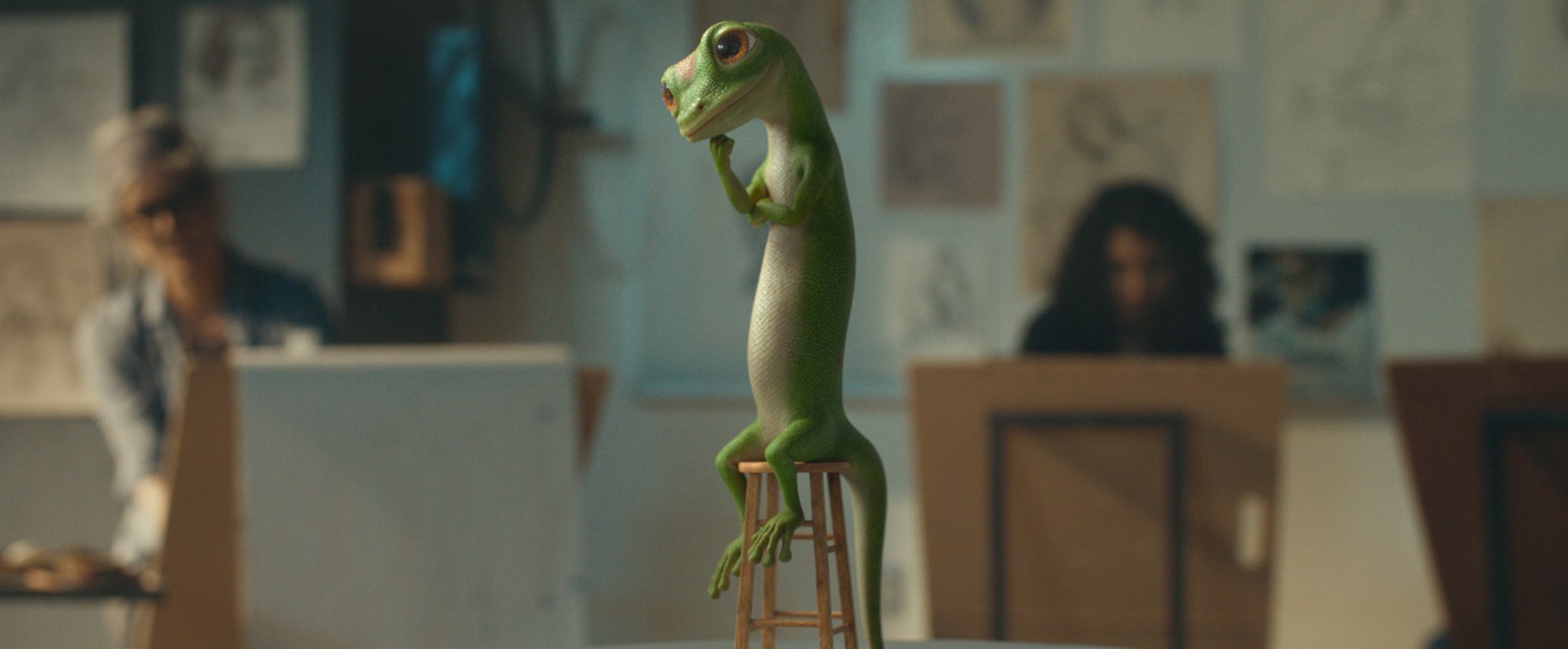 The CG animated GEICO gecko poses thoughtfully on a tiny stool in an art studio, surrounded by sketching artists and hanging drawings