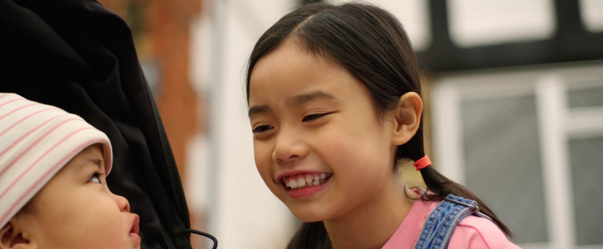A smiling young girl leans toward a baby in a stroller, sharing a warm, joyful moment outdoors.