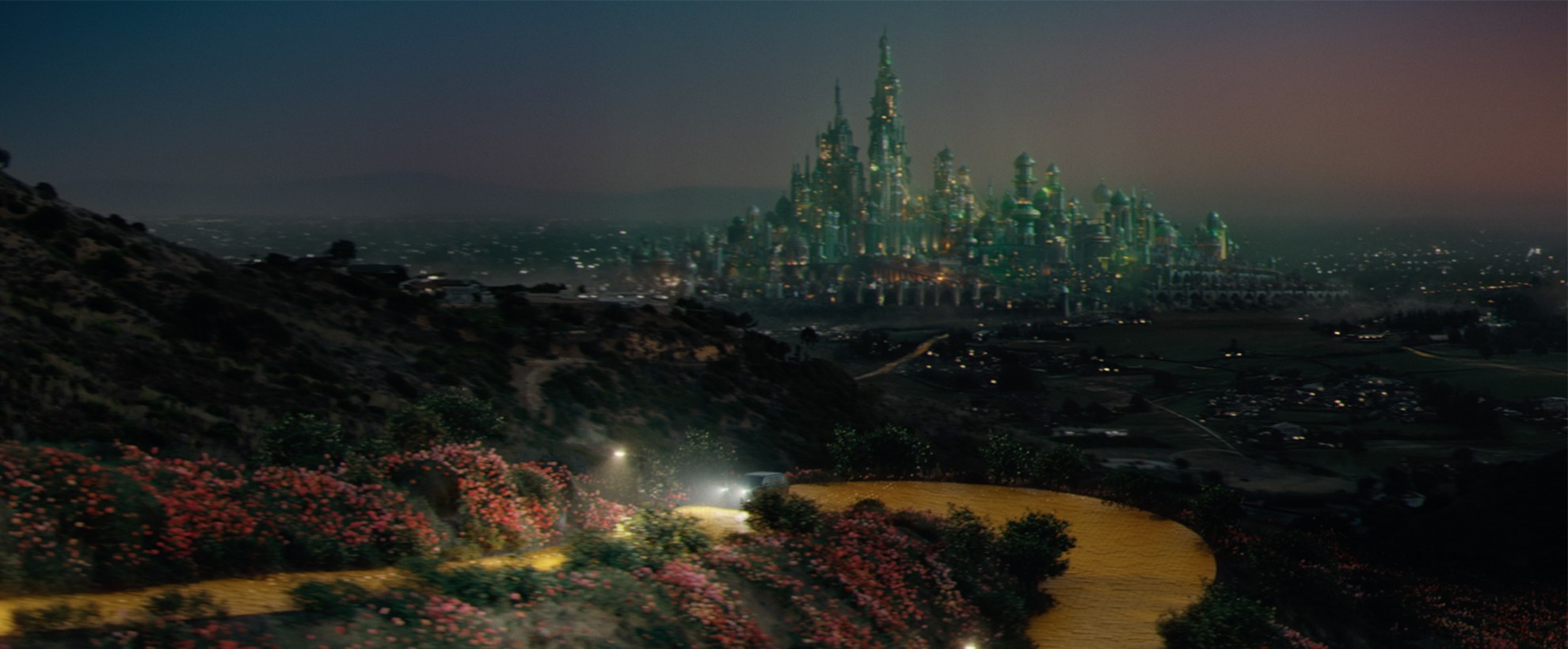 A glowing emerald city rises in the distance as a winding yellow road curves through flowered hills beneath twilight sky.