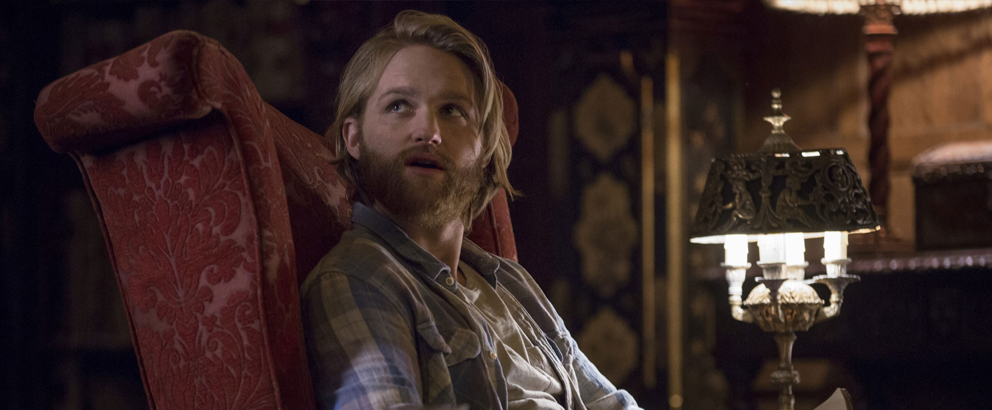 A blonde man sits in a red high-back armchair by a fireplace, looking over his shoulder