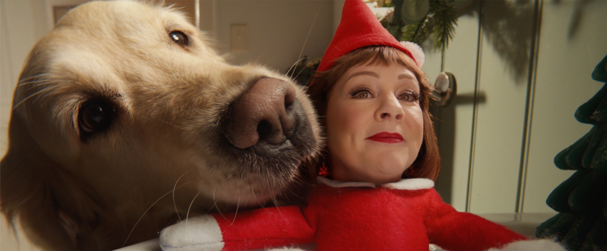 A golden retriever presses its nose toward the camera beside a small elf doll with Melissa McCarthy's comped face in an elf costume