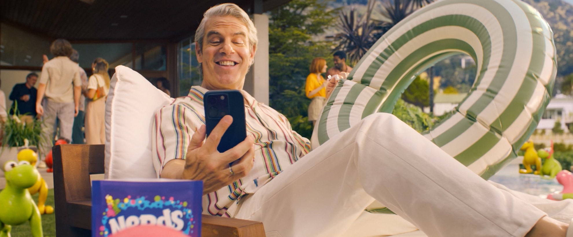 Andy Cohen sits on a pool lounge chair while answering a video call on his cell phone. He is holding an inflatable pool ring