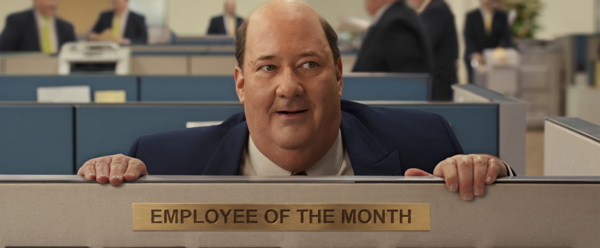 Actor Brian Baumgartner peers over a cubicle wall. There is an "Employee of the Month" placard on the wall