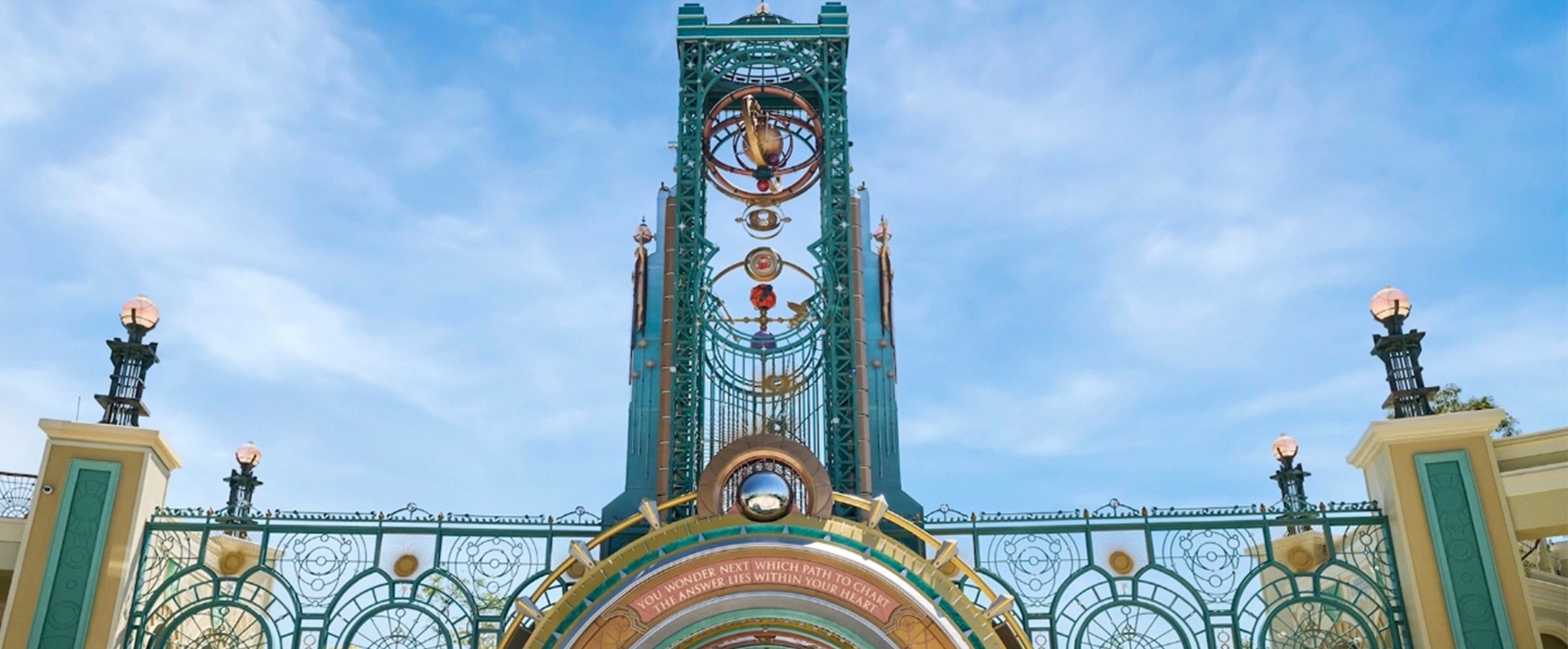 Ornate teal and gold theme park entrance with celestial clock tower, arched gateway, decorative ironwork, and cloudy blue sky backdrop.