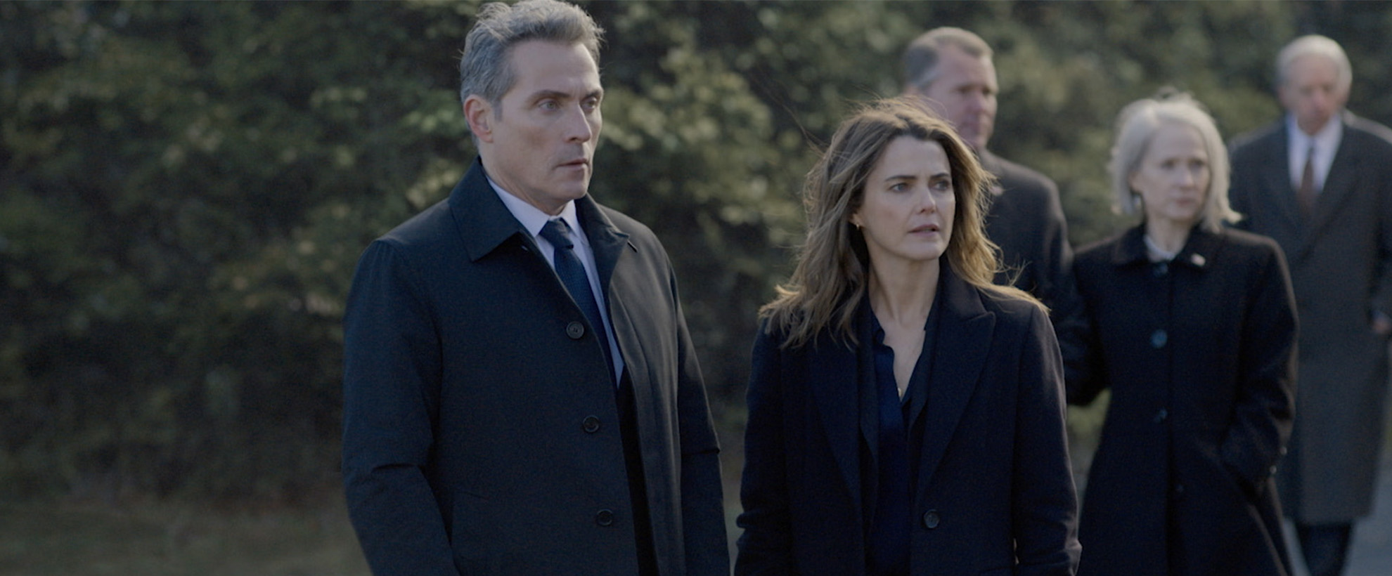 Kate Wyler (Keri Russell) and Hal Wyler (Rufus Sewell) in The Diplomat season 3. The two are in dark coats stand outdoors, appearing concerned as they look toward something unseen ahead together.