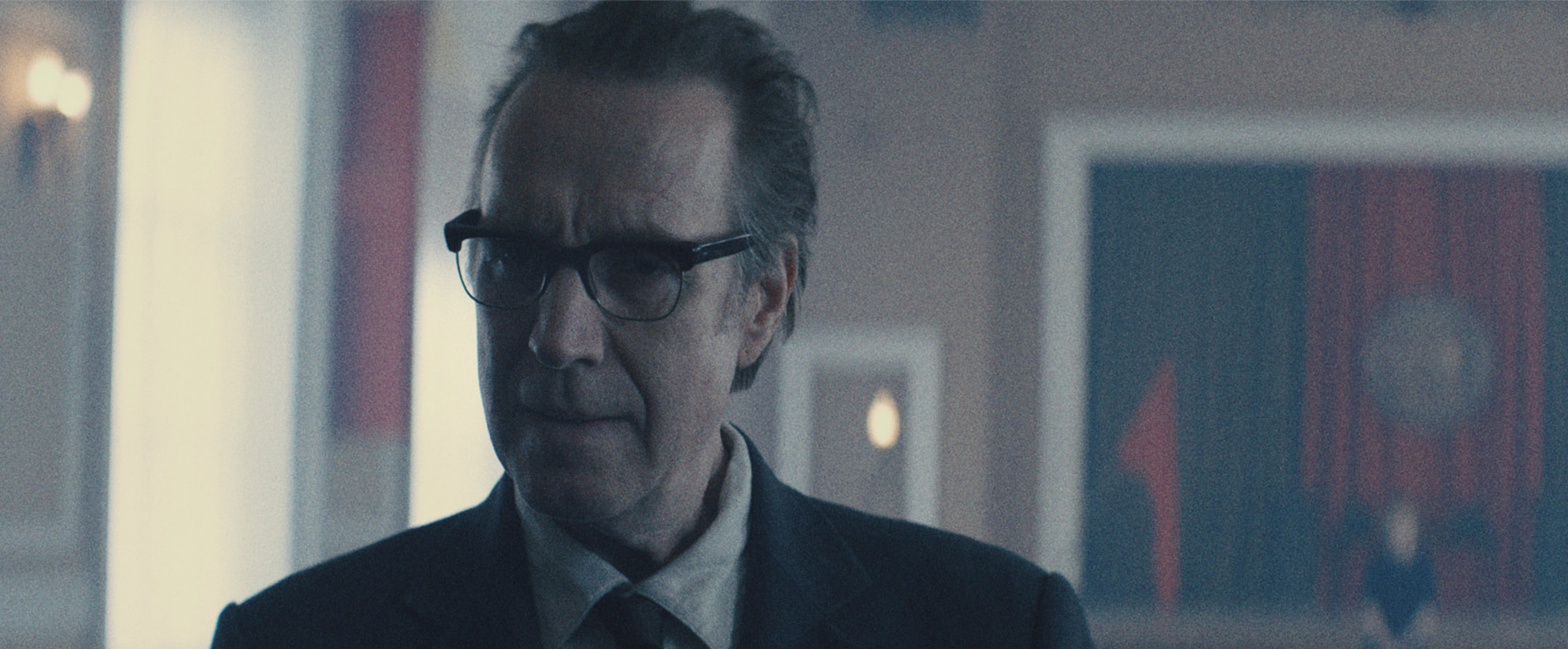 Actor Rhys Ifans in the tv show Star City, wearing glasses and a suit stands indoors, softly lit, with blurred artwork and warm lights in the background.