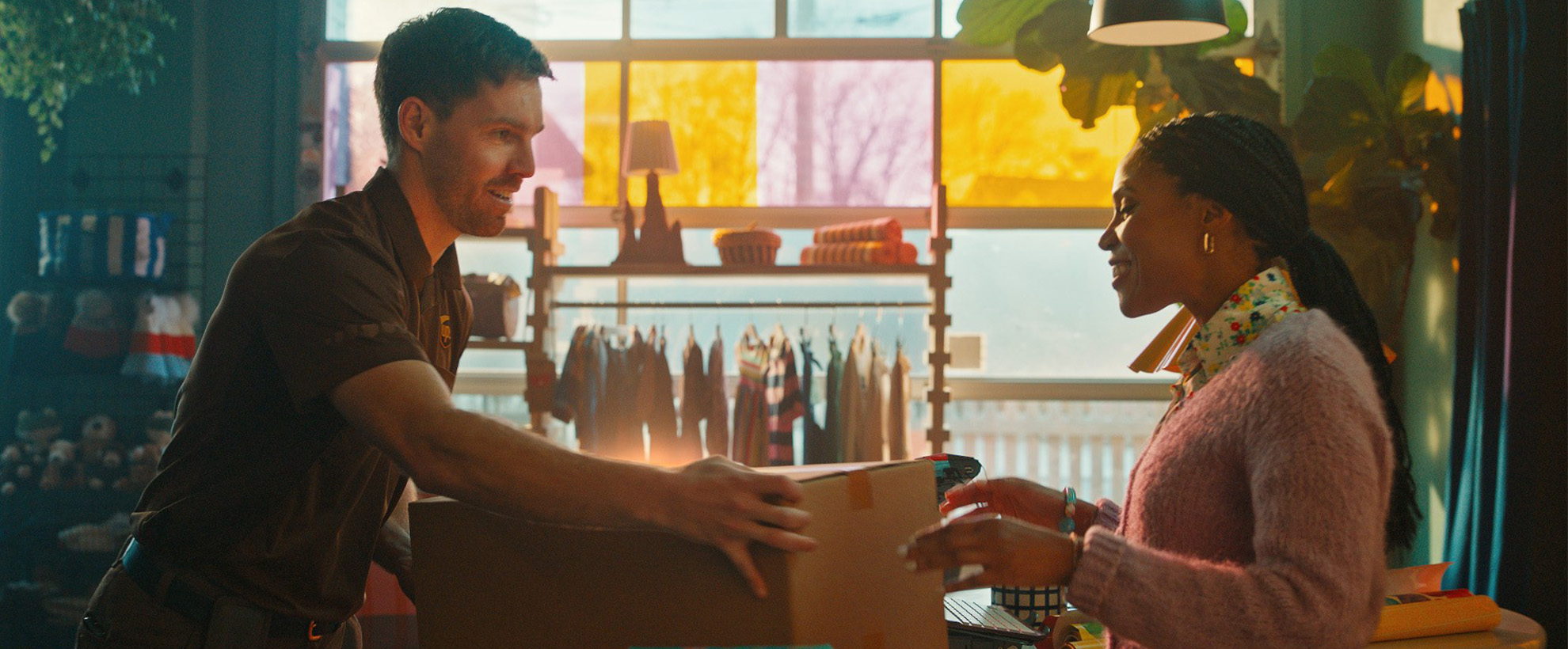 A UPS delivery worker hands a package to a smiling woman inside a colorful boutique with clothing racks and warm lighting.