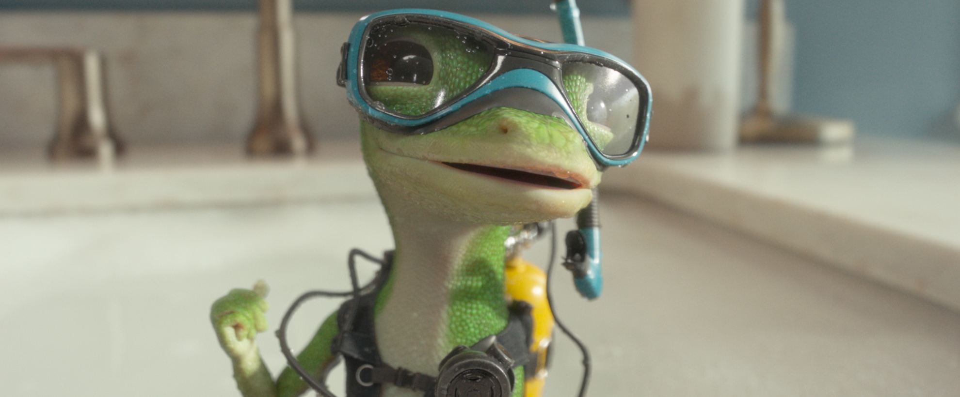 CG animated GEICO gecko wearing blue diving goggles and scuba gear stands in a bathroom sink.