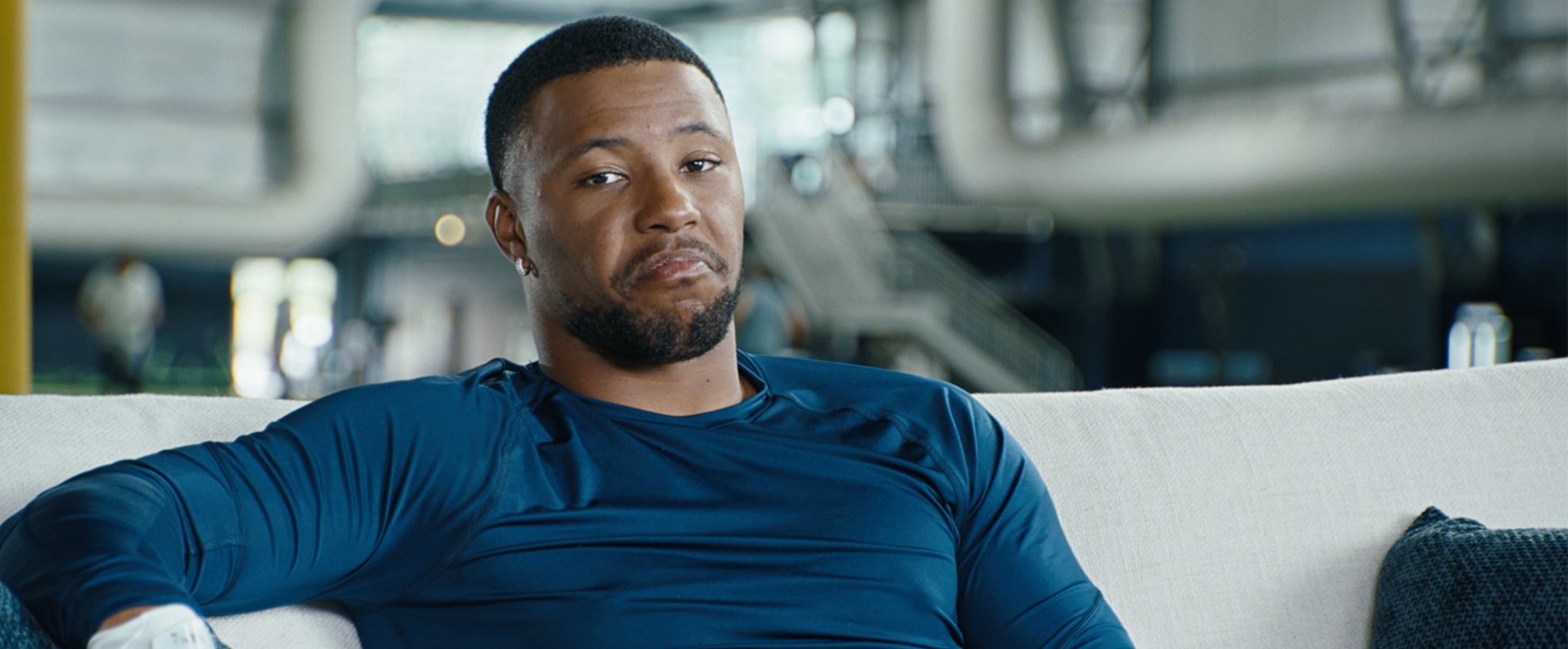 NFL star Saquon Barkley in a blue shirt sits on a couch, wearing white gloves, with an industrial interior behind him.