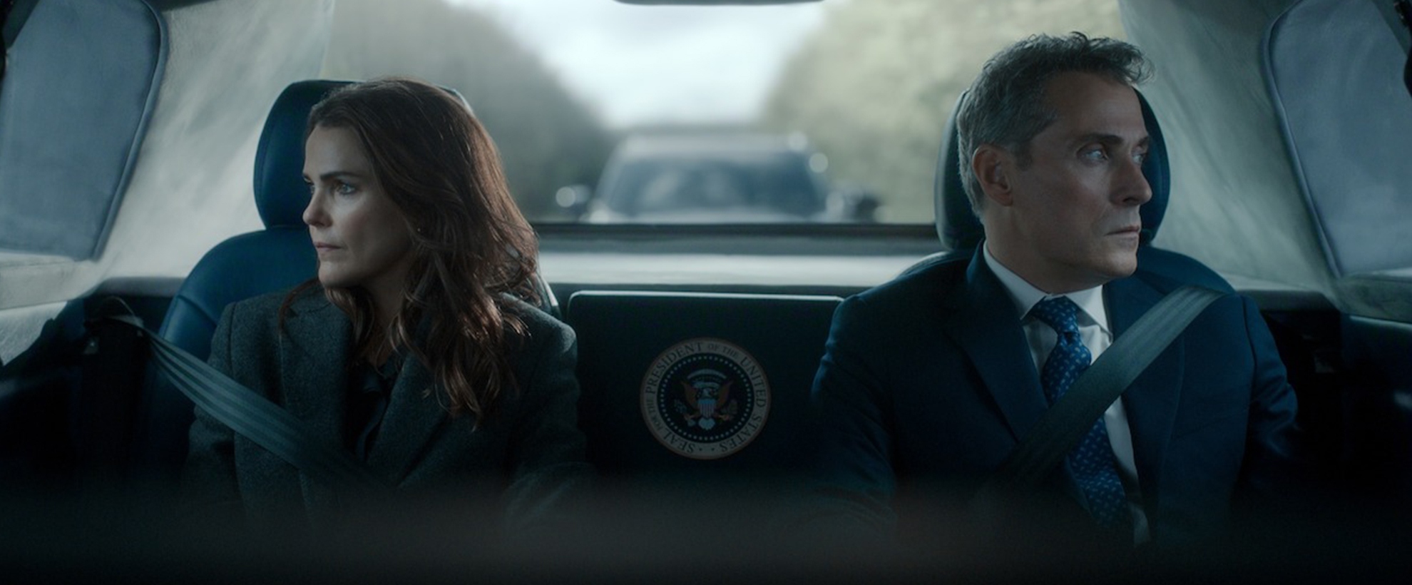 Keri Russell as Kate Wyler and Rufus Sewell as Hal Wyler in Netflix's The Diplomat Season 4 sit belted in the back of a government vehicle, gazing out opposite windows.