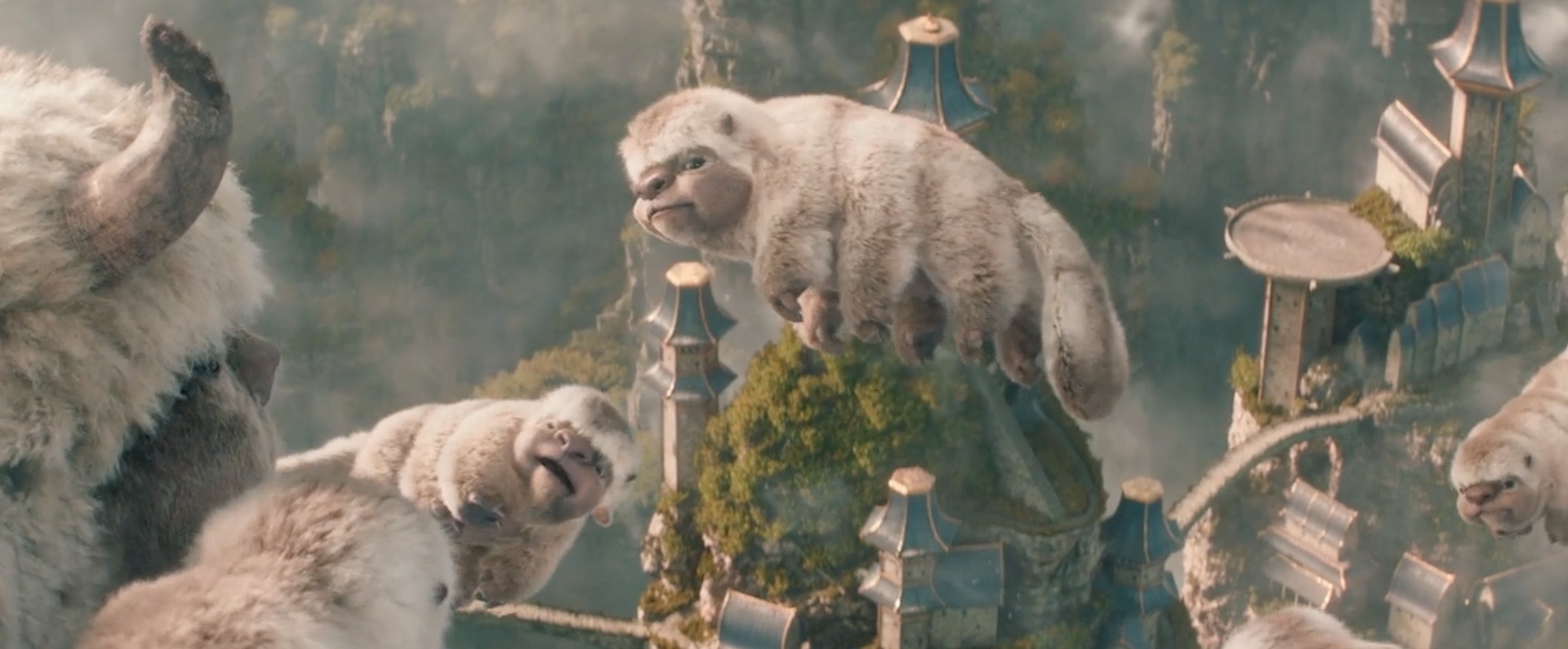 CG animated fluffy baby flying bison soar alongside an adult bison above a lush mountain landscape dotted with ornate temples and bridges.
