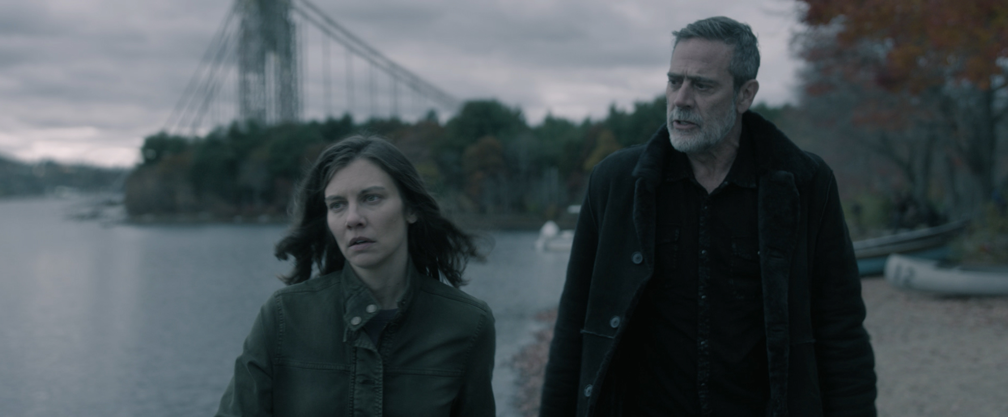 Lauren Cohan as Maggie Rhee and Jeffrey Dean Morgan as Negan Smith, looking somber and walking along a riverside beneath an overcast sky, with a broken suspension bridge and autumn trees behind them.