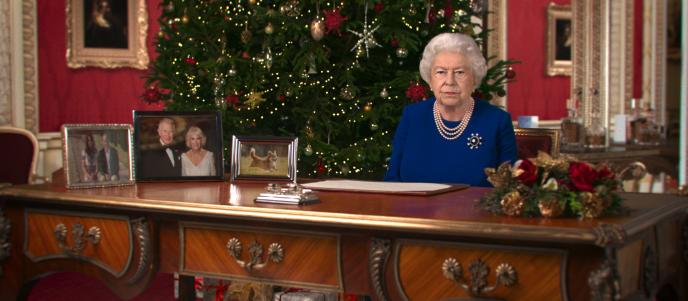 deepfake of queen elizabeth the second sitting behind a table. there is a large christmas tree behind her and the room is decorated festively 
