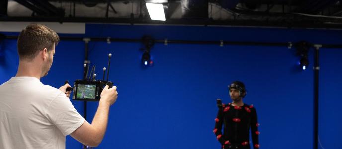 A man in a white t-shirt holds a small monitor, viewing a person in a motion capture suit against a blue screen background