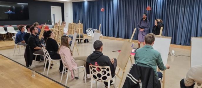  A group of people are seated with easels, observing a costumed model at the front of a well-lit room with a wooden floor and blue curtains.