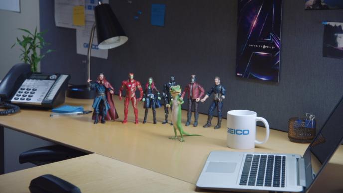 a startled geico gecko mascot standing in front of six avengers action figures. there is an avengers poster on the wall and a geico branded mug on the table