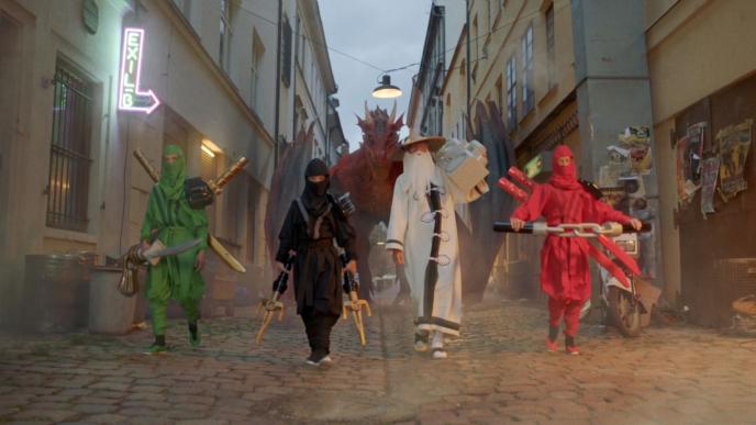 four characters in three ninja and one wizard costumes walk in front of a dragon as they are holding extra large toy weapons