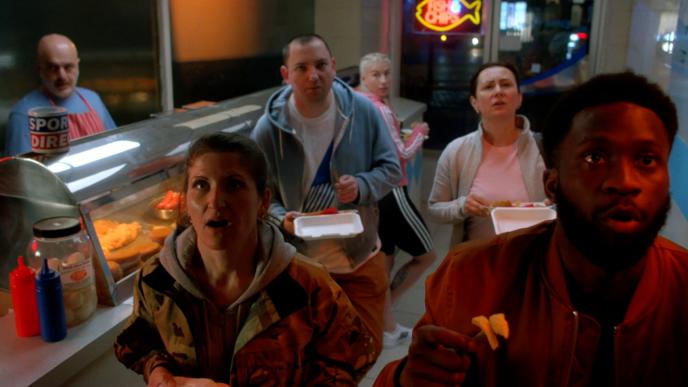 six people standing in a fish and chop shop looking up into a screen in shock and awe while holding food in their hands