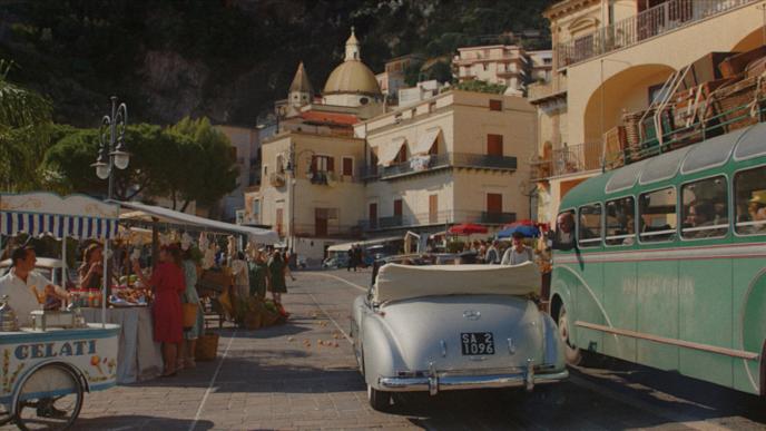 a food market and stalls at an italian coastal town. there is a 50s convertible car and a bus in the road
