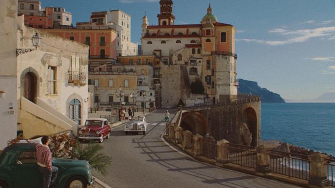 an italian coastal town set in the 50s. there are cars and people on the roadside. the sea and mountains are visible in the distance