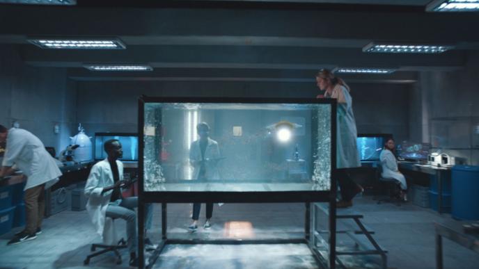 scientists in a dimly lit lab. there is an aquarium tank in the centre. there are three people looking into it. the scientist on the left is sitting down. the one on the right is standing on a step ladder peering into the tank from above. another one is behind the tank looking into it while holding a clipboard