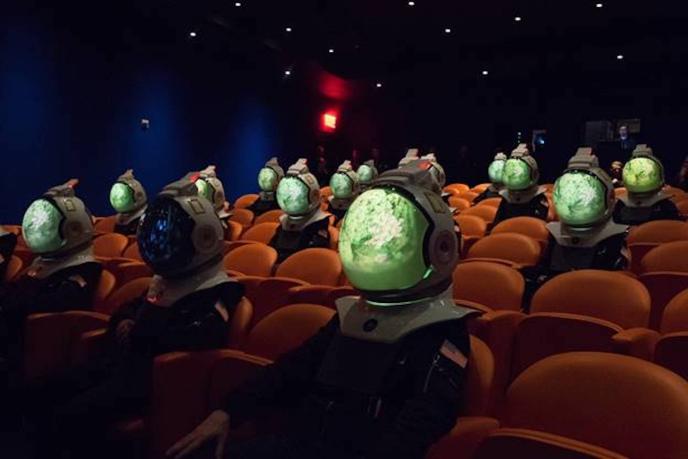 astronauts with nebula visuals in their helmets sitting in orange seats in a cinema