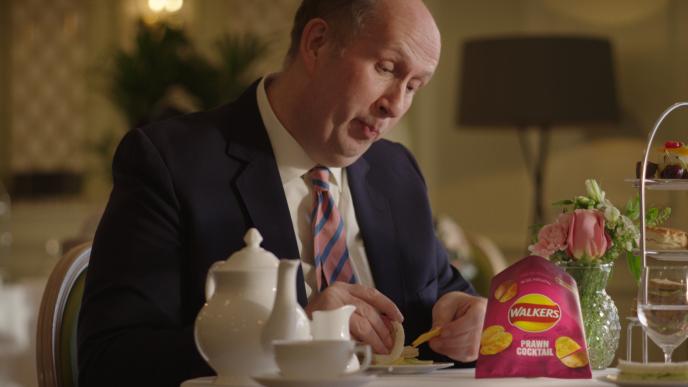 a person sitting at a table. there is a pot and cup of tea next to a bag of walkers crisps