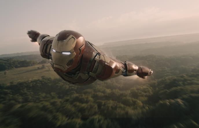 front side view of iron man flying in air with his arms to the sides of his body above forest greenery