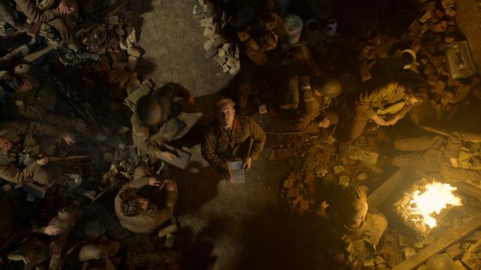 aerial shot of soldiers sitting and standing around rubble and campfire as a commander in the centre of the image is looking up directly into the camera