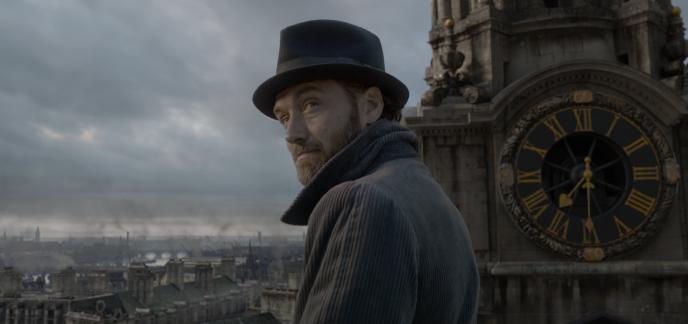 actor jude law as albus dumbledore looking back. he is wearing a fedora and grey jacket. he is standing next to a clock tower