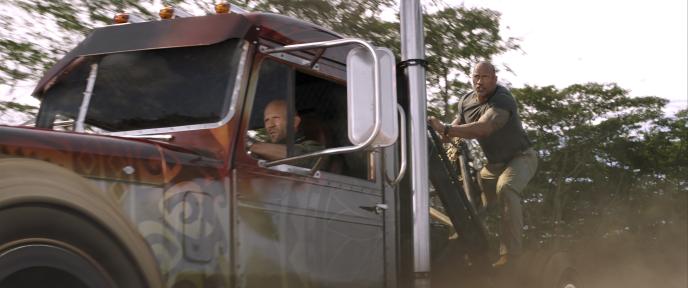 actor jason statham as deckard shaw driving a lorry and dwayne johnson as luke hobbs holding on the back of the cab side