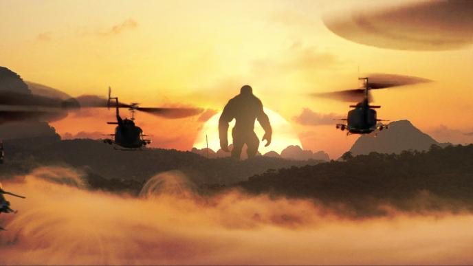 the silhoutte of king kong in front of a sunset as two helicopters approach it amongst mountainous terrain