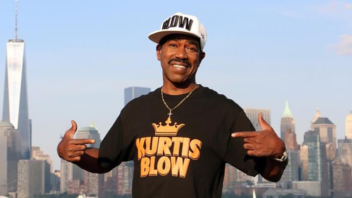 kurtis blow pointing at his kurtis blow branded t-shirt while smiling on a rooftop in front of the new york city skyline