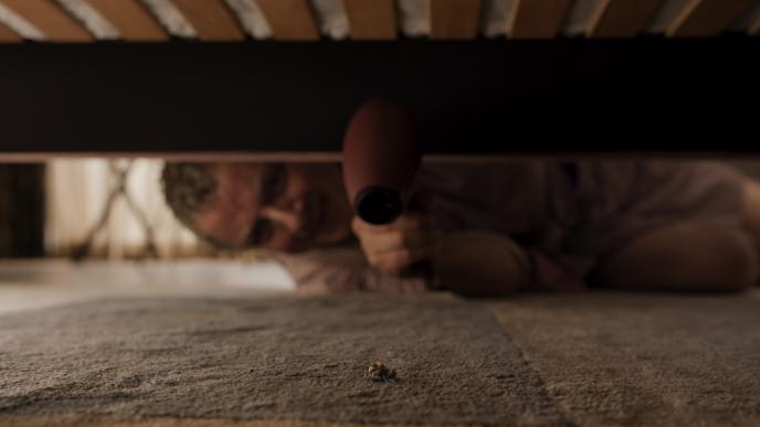 actor rowan atkinson laying on the floor looking at a bee under a bed while holding a hair dryer on it