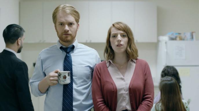 two people standing in an office kitchen. there are three people in the background