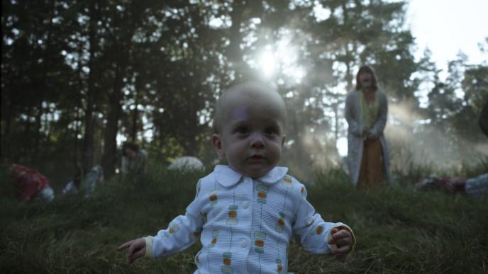 front view of a baby in a forest looking ahead as a woman stands yelling. there is a group of people on the grass in the background