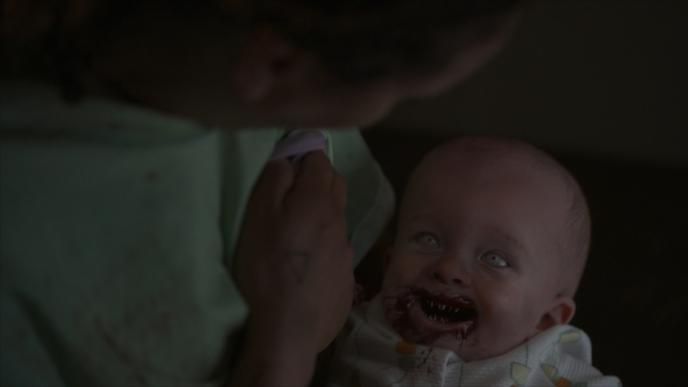 a posessed baby that has white eyes and sharp teeth inside a bloody mouth looking up at its mother