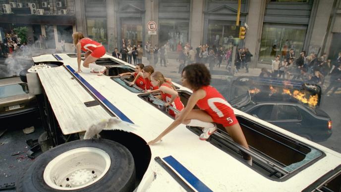 cheerleaders coming out of a bus that has fallen on its side as a car nearby is on fire and a crowd is rushing by