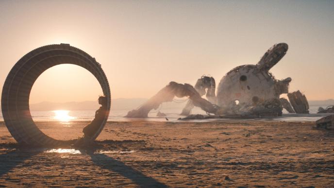person standing with their back against in a circular space amongst broken and abandoned amusement rides and attractions in a desert terrain