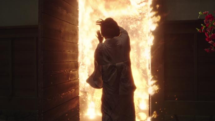 back view of a geisha wearing a white robe bracing herself as she stands in front of a door that is engulfed in flames