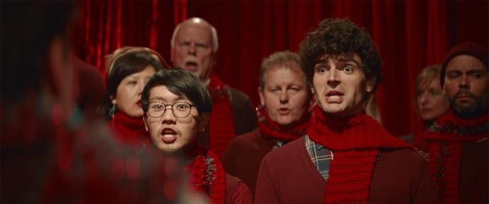 a christmas choir wearing red scarves and jumpers singing hastily while looking confused