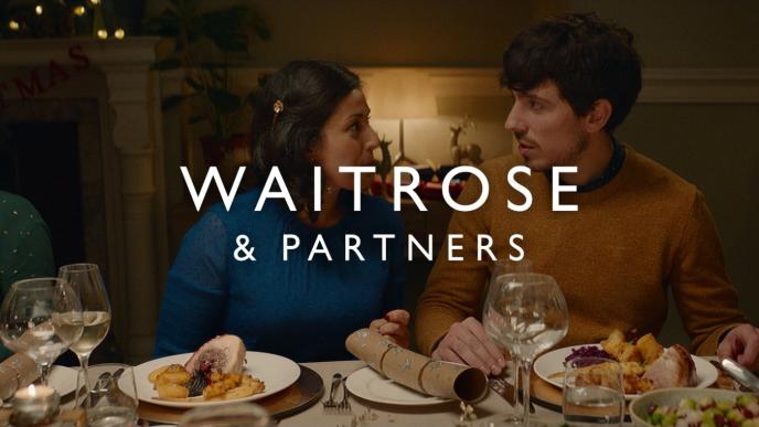 a couple sitting at a christmas dinner table looking at each other. there is 'waitrose & partners' text in the centre of the image