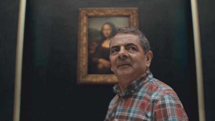 actor rowan atkinson with a silly smile in front of the mona lisa painting