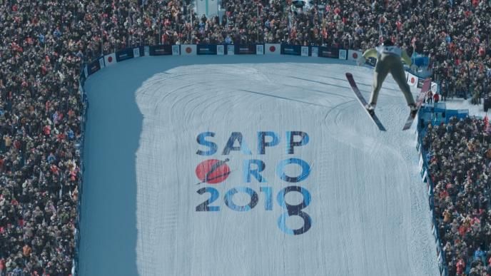 a large crowd surrounds a ski ring as an olympic skiier descends from the sky
